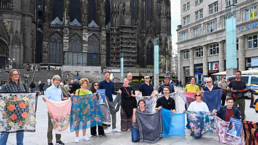 Anl&auml;sslich des 30. Jahrestages des Genozids von Srebrenica halten Mitarbeitende von Pro Peace T&uuml;cher und Schals vor dem K&ouml;lner Dom empor