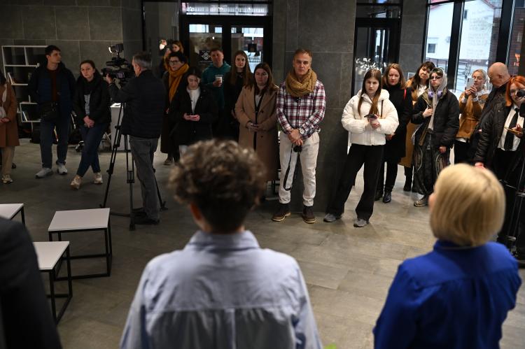 A group of people stands in a gallery space listening to speakers during the exhibition opening
