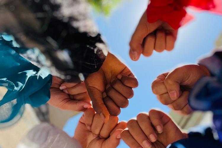 People stand in a circle and stretch their hands toward the center. They wear scarves around their wrists.