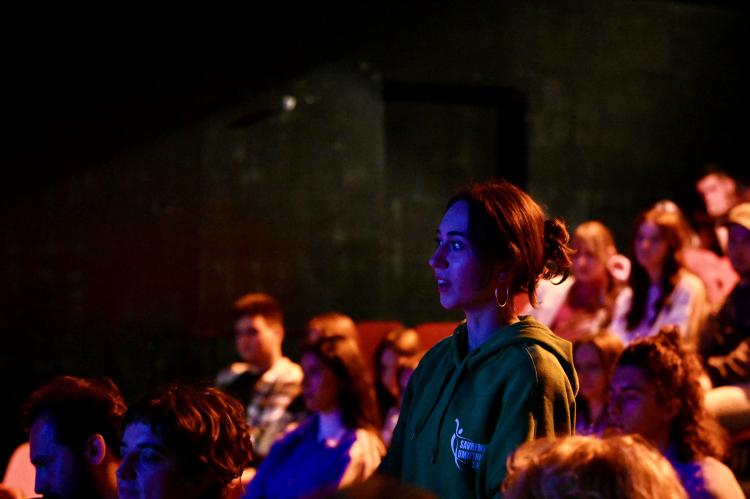 A girl stands surrounded by an audience and speaks to the people on stage.