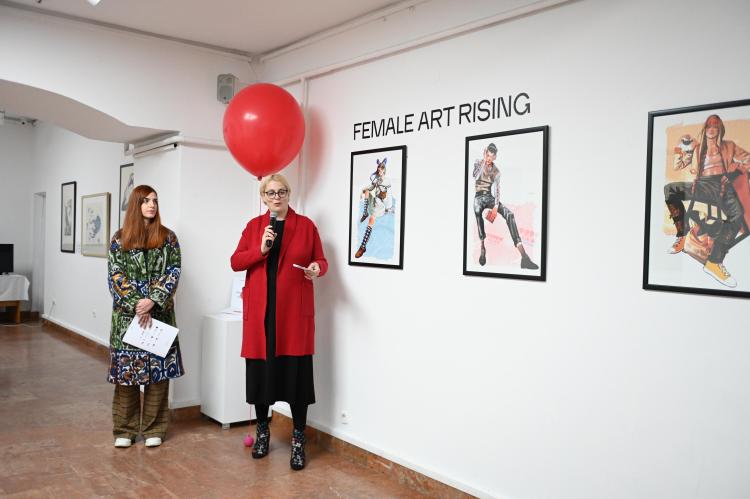 A speaker stands next to a red ballon and some of the artworks and speaks to an audience.