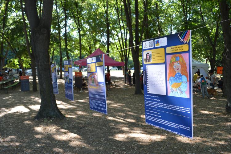 Drei Ausstellungsplakate der Ausstellung "Gesichter ukrainischer Träumer" werden im Schatten mehrerer Bäumer präsentiert.