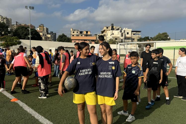Zwei Mädchen in Fußballtrikots stehen am Rande einer Gruppe von Jugendlichen und haben sich ihre Arme um die Schultern gelegt.