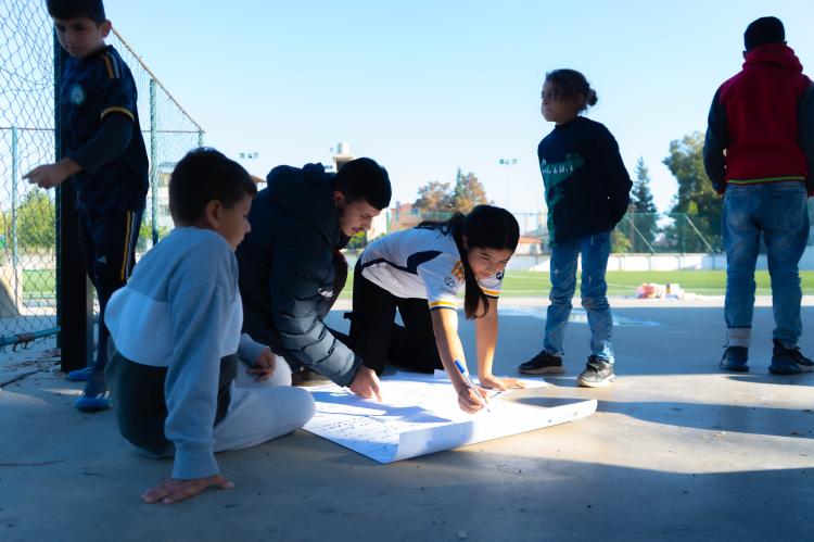 A group of children are sitting on the concrete floor of a sports facility and painting a poster.