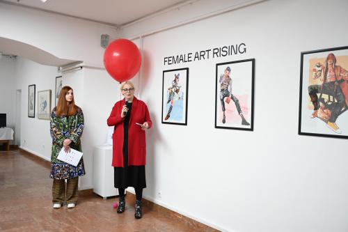 Eine Rednerin steht vor einem roten Ballon und einigen Kunstwerken und spricht zum Publikum.