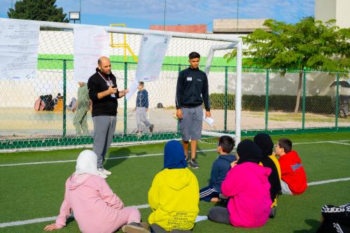 A group of children are sitting on a soccer field. Two adult men are standing in front of them and talking to them.