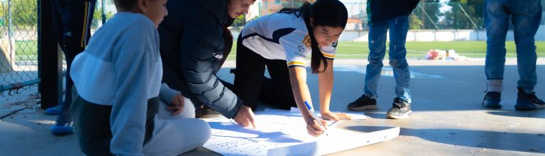 Eine Gruppe von Kindern sitzt auf dem Betonboden einer Sportanlage und malt ein Plakat.