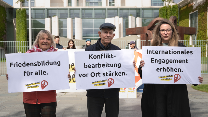 Drei Personen halten vor dem Kanzleramt in Berlin die zentralen Forderungen der Kampagne "Friedensstandort Deutschland" in die Höhe Friedensstandort Deutschland: Protestaktion von Pro Peace vor dem Kanzleramt