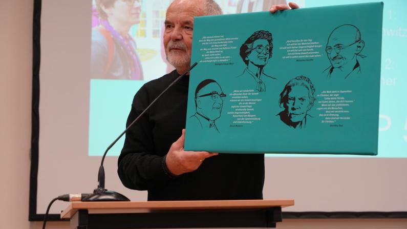 Gerold König spricht vor Publikum und hält ein Plakat mit Friedensaktivist*innen in die Höhe.