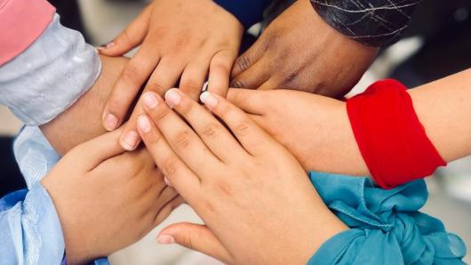 People stand in a circle and stretch their hands toward the center. They wear scarves around their wrists.
