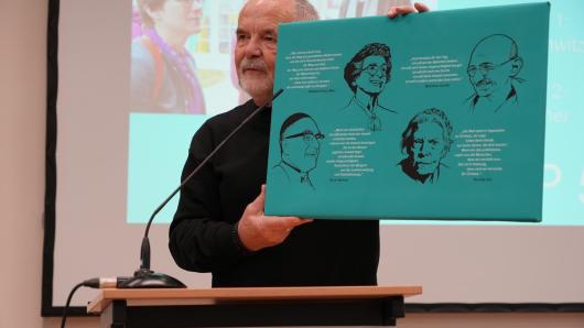 Gerold K&ouml;nig spricht vor Publikum und h&auml;lt ein Plakat mit Friedensaktivist*innen in die H&ouml;he.