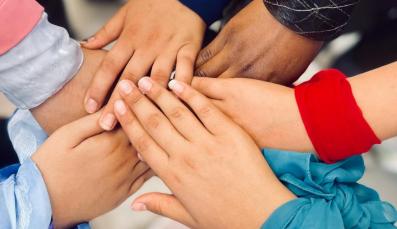 People stand in a circle and stretch their hands toward the center. They wear scarves around their wrists.