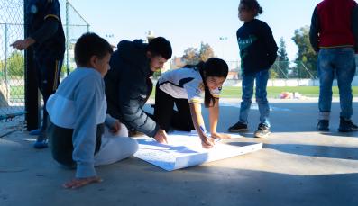 Eine Gruppe von Kindern sitzt auf dem Betonboden einer Sportanlage und malt ein Plakat.