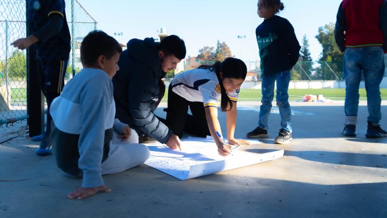 Eine Gruppe von Kindern sitzt auf dem Betonboden einer Sportanlage und malt ein Plakat.